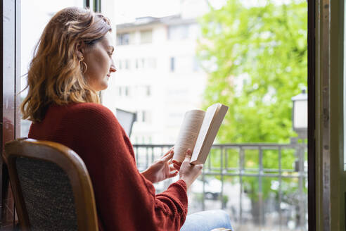 Junge Frau liest ein Buch an der Balkontür - FBAF00801