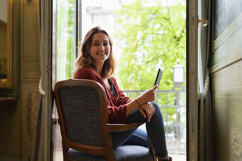 Lächelnde junge Frau an der Balkontür sitzend mit Mobiltelefon - FBAF00802