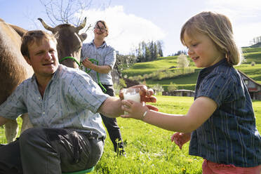 Farmer giving his daughter a glass of fresh milk from a cow on pasture - FBAF00843