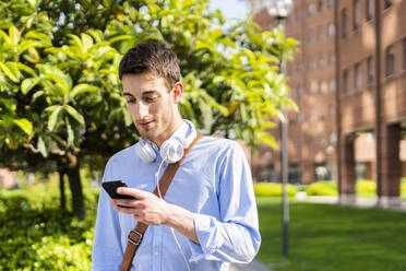 Young man using smartphone, headphones around neck - GIOF06513