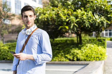 Young man holding smartphone, headphones around neck - GIOF06514