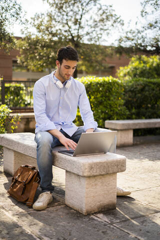 Junger Mann mit Laptop, Kopfhörer um den Hals, auf einer Bank sitzend, lizenzfreies Stockfoto
