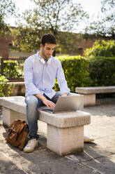 Young man using laptop, headphones around neck, sitting on bench - GIOF06517