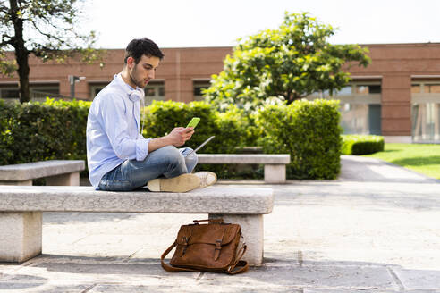 Junger Mann mit Laptop und Smartphone, Kopfhörer um den Hals, auf einer Bank sitzend - GIOF06518