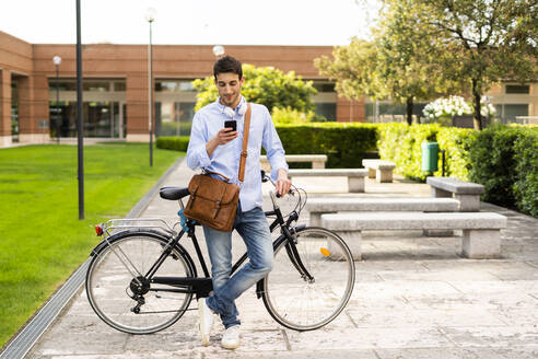 Junger Mann mit Fahrrad und Smartphone, Kopfhörer um den Hals in der Stadt - GIOF06521