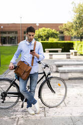 Young man with bicycle using smartphone, headphones around neck in the city - GIOF06522