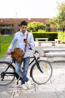 Junger Mann mit Fahrrad und Smartphone, Kopfhörer um den Hals in der Stadt - GIOF06522
