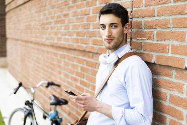 Young man with bicycle using smartphone, headphones around neck - GIOF06523