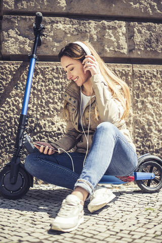 Happy woman sitting on E-Scooter listening music with headphones and smartphone stock photo