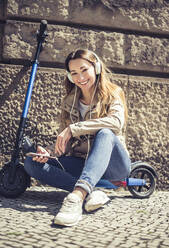 Porträt einer glücklichen Frau, die auf einem E-Scooter sitzt und mit Kopfhörern und Smartphone Musik hört - BFRF02041