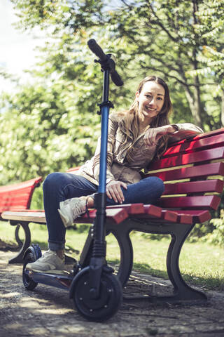 Porträt einer lächelnden Frau mit E-Scooter, die sich auf einer Bank ausruht, lizenzfreies Stockfoto