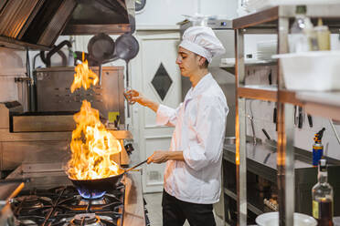 Junior chef with pan of flames in traditional spanish restaurant kitchen - LJF00198