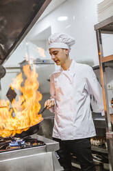 Junior chef with pan of flames in traditional spanish restaurant kitchen - LJF00200