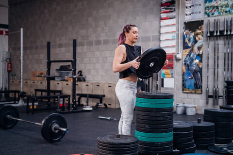 Junge Frau legt eine Hantelscheibe auf ein Gestell im Fitnessstudio, lizenzfreies Stockfoto