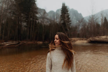 Young woman on riverbank shaking long brown hair, Yosemite Village, California, USA - ISF22113