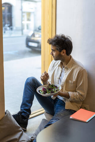 Mann isst einen Salat, sitzt am Fenster in einem Restaurant und schaut hinaus, lizenzfreies Stockfoto
