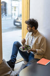 Mann isst einen Salat, sitzt am Fenster in einem Restaurant und schaut hinaus - AFVF03552