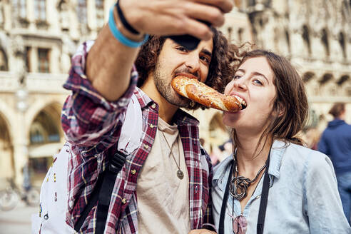 Young couple taking a selfie with brezel in the mouth, Munich, Germany - SUF00582