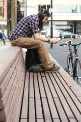 Seitenansicht eines bärtigen Mannes im mittleren Erwachsenenalter, der auf einer Bank in der Stadt sitzt und Zeitung liest - IGGF01189