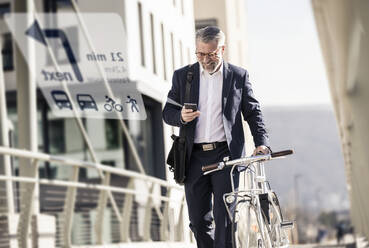 Lächelnder reifer Geschäftsmann mit Fahrrad, der ein Mobiltelefon zur Navigation in der Stadt benutzt - UUF17913