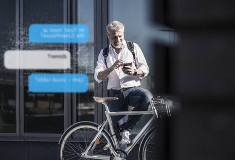 Lächelnder reifer Geschäftsmann mit Fahrrad, der ein Mobiltelefon für Nachrichten in der Stadt benutzt - UUF17916