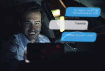 Lächelnder Geschäftsmann, der ein Handy benutzt, um mir nachts im Auto Nachrichten zu schicken - UUF17923