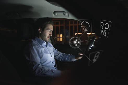 Geschäftsmann, der nachts ein Gerät im Auto benutzt, umgeben von Internet-Symbolen - UUF17925