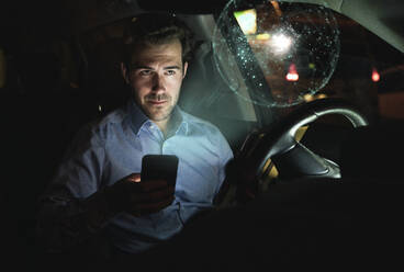 Businessman using smartphone in car at night surrounded by virtual shining globe - UUF17927