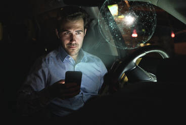 Geschäftsmann, der nachts im Auto ein Smartphone benutzt, umgeben von einer virtuellen leuchtenden Weltkugel - UUF17927