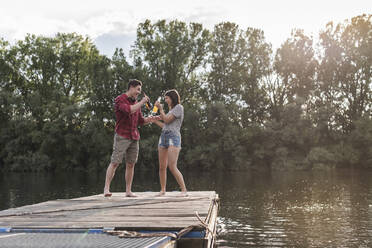 Happy young couple having a drink on jetty at a remote lake - UUF17935