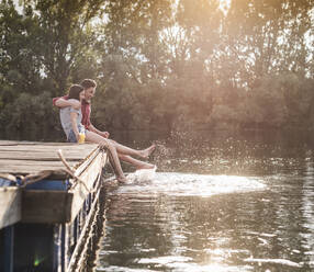 Happy young couple having a drink and splashing with water on jetty at a remote lake - UUF17937