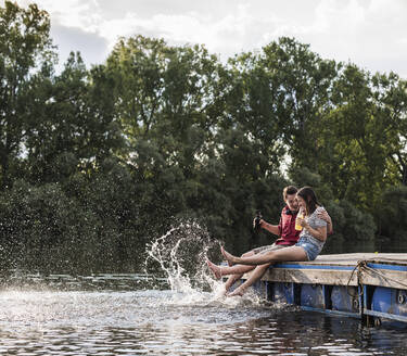 Junges Paar trinkt und plantscht auf einem Steg an einem abgelegenen See - UUF17940