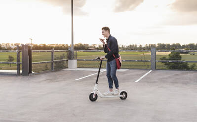 Glücklicher junger Mann mit Handy und Elektroroller auf einem Parkdeck - UUF17953