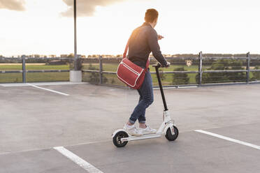 Junger Mann mit Handy und Elektroroller auf Parkdeck bei Sonnenuntergang - UUF17955