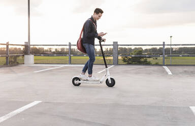 Happy young man with cell phone and electric scooter on parking deck at sunset - UUF17956