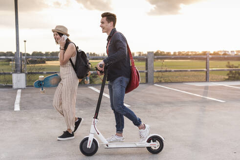 Glückliches junges Paar mit Handy, Longboard und Elektroroller auf dem Parkdeck - UUF17959