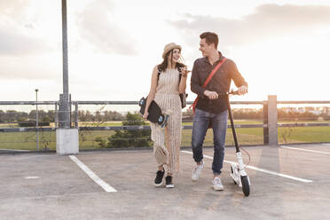 Glückliches junges Paar mit Handy, Longboard und Elektroroller auf dem Parkdeck - UUF17960