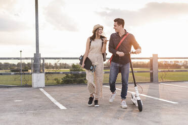 Happy young couple with cell phone, longboard and electric scooter on parking deck - UUF17960