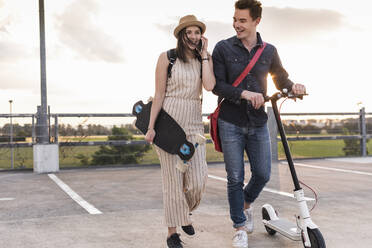 Happy young couple with cell phone, longboard and electric scooter on parking deck - UUF17961