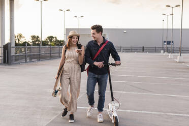 Happy young couple with cell phone, longboard and electric scooter on parking deck - UUF17962