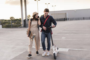 Happy young couple with cell phone, longboard and electric scooter on parking deck - UUF17963