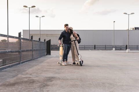 Junges Paar mit Longboard und Elektroroller umarmt sich auf dem Parkdeck, lizenzfreies Stockfoto