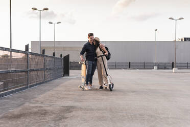 Young couple with longboard and electric scooter hugging on parking deck - UUF17965