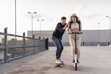 Young man and woman riding on longboard and electric scooter on parking deck - UUF17966