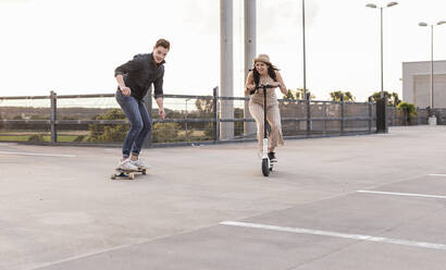 Junger Mann und Frau fahren auf Longboard und Elektroroller auf einem Parkdeck - UUF17967