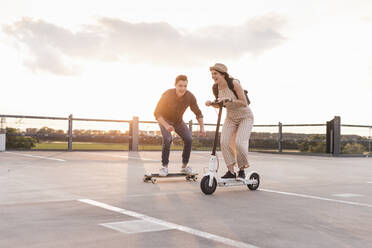 Young man and woman riding on longboard and electric scooter on parking deck at sunset - UUF17968
