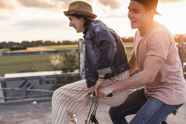 Happy young couple together on a bicycle on parking deck at sunset - UUF17977
