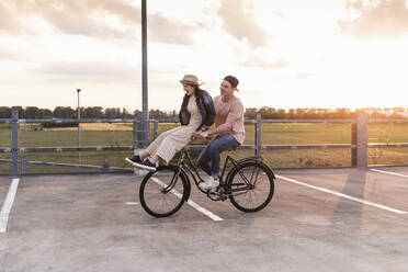 Happy young couple together on a bicycle on parking deck at sunset - UUF17979