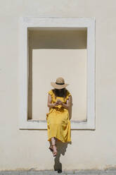 Frau mit Hut, die ein Mobiltelefon benutzt, während sie auf einem Bauwerk in der Stadt sitzt - AHSF00572