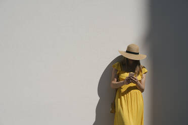 Weiblicher Tourist mit Hut, der sein Smartphone benutzt, während er in der Stadt an der Wand steht - AHSF00575